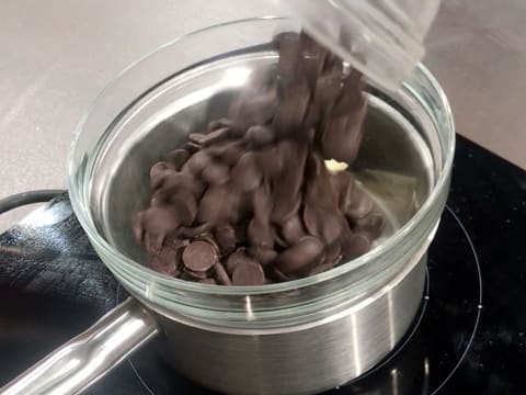 Ajout des pistoles de chocolat noir sur le beurre dans le saladier en verre qui est placé sur une casserole contenant de l'eau