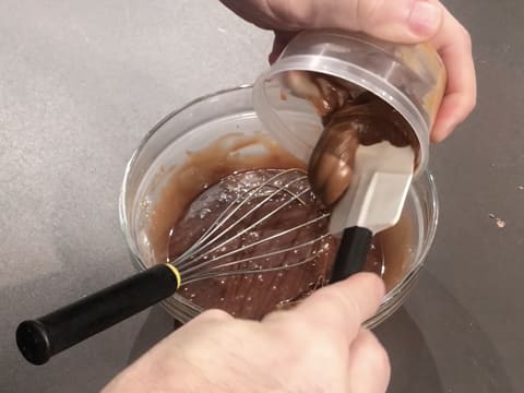 Ajout du praliné amandes noisettes dans la préparation chocolatée qui se trouve dans le saladier en verre posé sur le plan de travail