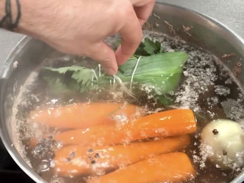 Ajout d'un bouquet garni dans l'eau frémissante dans le grand faitout qui contient les morceaux de viande, quatre carottes, un oignon et les grains de poivre