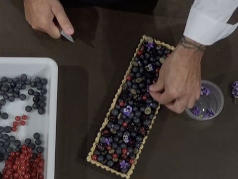 Des fleurs comestibles violettes et de petite taille sont disposées sur la tarte, insérées entre les fruits