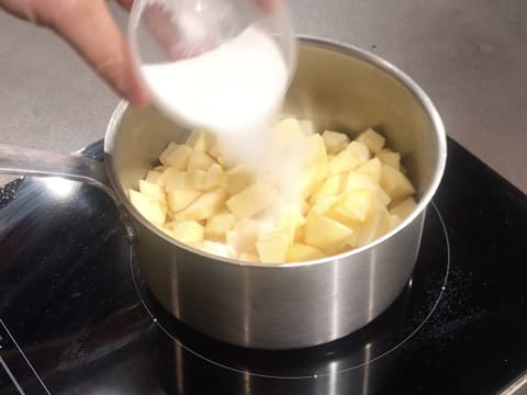 Du sucre en poudre est versé dans la casserole qui contient les morceaux de pommes et qui est placée sur une plaque de cuisson