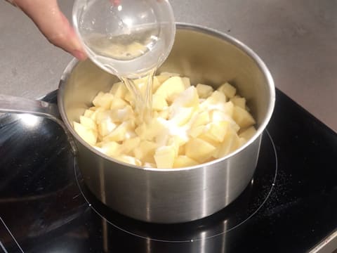 Ajout de l'eau sur le sucre et les morceaux de pommes dans la casserole qui est placée sur une plaque de cuisson