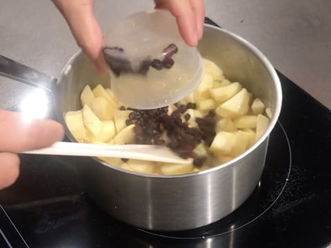 Ajout des raisins secs sur les pommes et le sucre dans la casserole qui contient les morceaux de pommes et qui est placée sur une plaque de cuisson