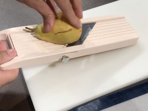 Une poire est émincée à l'aide d'une mandoline, au-dessus d'une planche à découper qui est posée sur le plan de travail