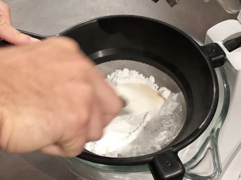 La spatule maryse est passée sur le sucre glace dans la passoire tamis qui est posée sur la cuve du batteur électrique