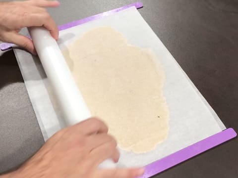 La pâte à streusel amande est abaissée de manière horizontale au rouleau à pâtisserie, entre deux feuilles de papier sulfurisé et entre deux réglettes à niveler violettes