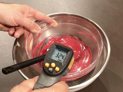 Prise de la température du chocolat inspiration framboise dans le bol en verre qui trempe dans le cul de poule rempli d'eau, à l'aide d'un thermomètre à visée laser qui affiche 32,4°C