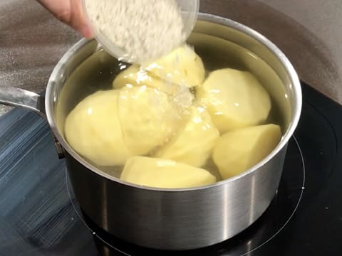 Ajout de gros sel dans la casserole contenant l'eau et les pommes de terre qui est placée sur une plaque de cuisson