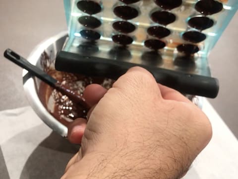 Le moule à chocolat demi sphère est raclé à la surface à l'aide de la spatule à chocolat, au dessus du cul de poule qui contient le chocolat noir fondu et tempéré