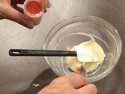 La pointe d'un couteau de colorant en poudre orange est ajoutée sur le chocolat blanc fondu dans le saladier en verre