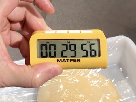 Vision d'un minuteur qui affiche 29 minutes et 56 secondes en premier plan, avec le bac alimentaire contenant la pâte à brioche qui est filmé, en arrière plan