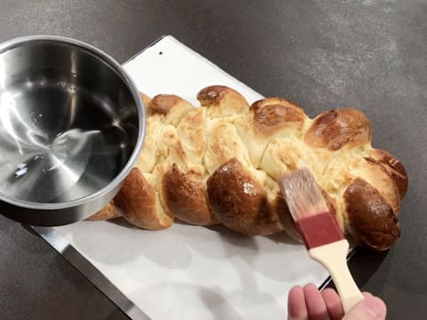 À l'aide d'un pinceau pâtissier, le pain brioché tressé est nappé de sirop de lustrage qui se trouve dans une petite casserole