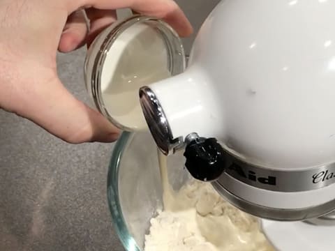 La crème liquide est versée dans la cuve du batteur sur les ingrédients qui sont en train d'être mélangés avec l'accessoire crochet