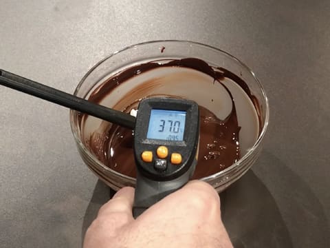 Prise de la température du chocolat noir fondu dans le saladier en verre, à l'aide d'un thermomètre à visée laser qui affiche 37,0°C