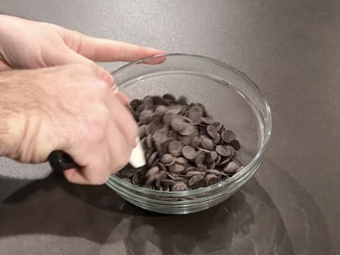 Les pistoles de chocolat noir sont mélangées avec une spatule type maryse dans un saladier en verre qui est posé sur le plan de travail