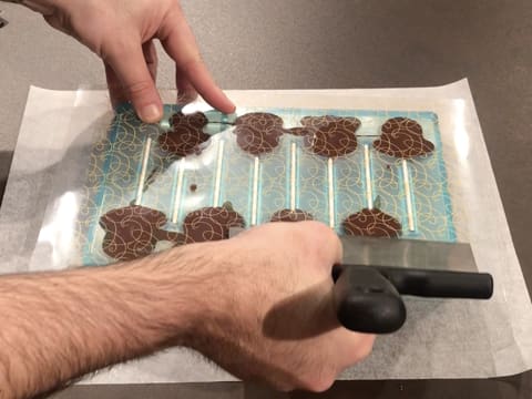 La spatule à chocolat est passée sur la feuille transfert qui recouvre le moule sucettes dont les cavités sont remplies de chocolat noir tempéré