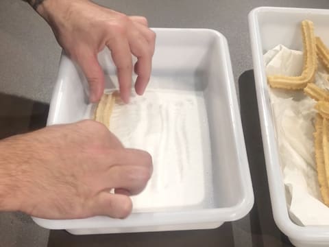 Un churros égoutté est passé dans le sucre en poudre qui est dans un bac alimentaire