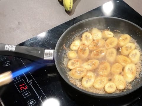 La flamme d'un chalumeau de cuisine est présentée sur le rebord de la poêle qui contient les rondelles de banane dans le caramel bond et le rhum brun