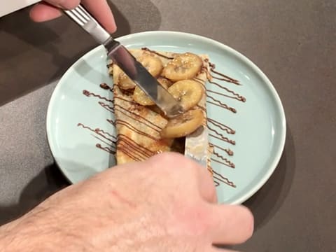 Des rondelles de banane cuite sont disposées sur la crêpe décorée de pâte à tartiner, à l'aide de deux petites spatules métalliques coudées