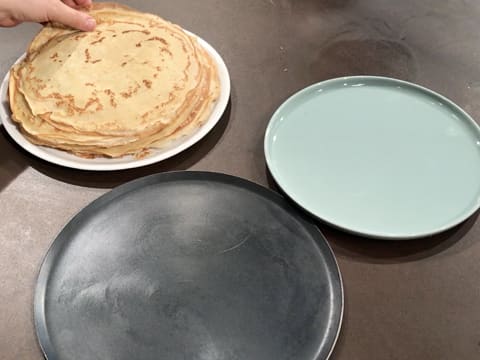 En arrière plan, les crêpes sont posées sur une assiette et en premier plan vision d'une plaque à pâtisserie ronde et une seconde assiette est placée sur la droite, sur le plan de travail