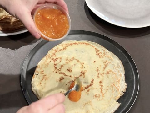 Une cuillère à café de confiture d'abricot est déposée sur la crêpe qui est placée sur une plaque à pâtisserie ronde