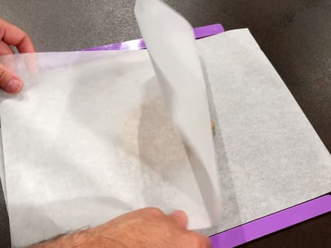 La pâte à streusel noisette qui est sur une feuille de papier sulfurisé posée sur le plan de travail, est recouverte d'une seconde feuille de papier sulfurisé et entourée de deux réglettes à niveler