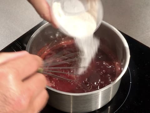 Ajout du mélange pectine et sucre dans la casserole qui contient la préparation à base de purées de fraise et de griotte qui est en train de cuire et d'être mélangée au fouet