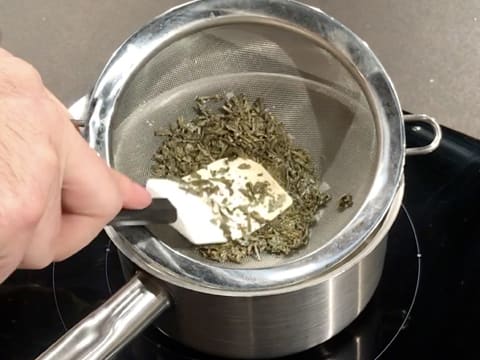 Les feuilles de thé au jasmin sont écrasées avec la spatule maryse dans le fond de la passoire fine au-dessus de la casserole sur la plaque de cuisson