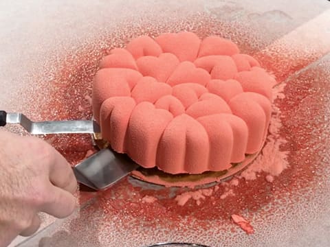 L'entremets coloré en rose est soulevé à l'aide de deux spatules métalliques coudées