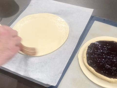 De l'eau est appliquée au pinceau à pâtisserie sur tout le pourtour du second disque de pâte feuilletée inversée qui se trouve sur une feuille de papier sulfurisé