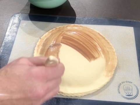 La dorure cacao est appliquée au pinceau pâtissier sur toute la surface de la galette des rois qui est sur une plaque à pâtisserie recouverte d'un tapis de cuisson en silicone