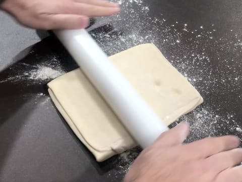 Le pâton rectangulaire qui a deux marques, est étalé sur le plan de travail fleuré, à l'aide du rouleau à pâtisserie