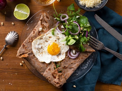 Obtention de la galette de sarrasin farcie et son œuf à cheval, qui est présentée sur assiette de service, et accompagnée d'un peu de salade et de fines lamelles d'oignon rouge