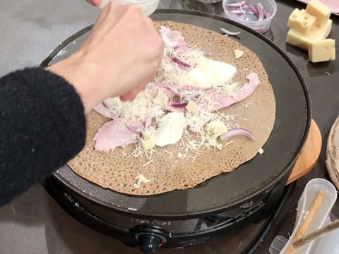 De l'emmental râpé est parsemé sur les autres ingrédients qui garnissent la galette qui cuit sur la crêpière