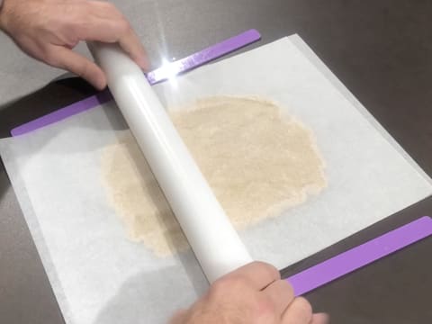 La pâte à streusel amande est recouverte d'une feuille de papier sulfurisé et deux réglettes à niveler violettes sont au premier plan