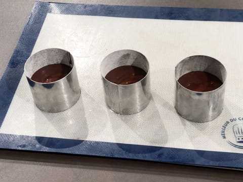 Trois cercles à vacherin en inox qui sont placés sur une plaque à pâtisserie recouverte d'un tapis de cuisson en silicone, sont remplis aux trois quarts de préparation chocolatée