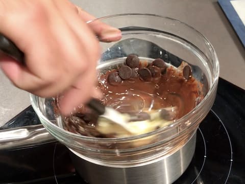 Les pistoles de chocolat et le beurre qui sont dans le saladier sur la casserole, sont en train de fondre tout en étant mélangés avec une spatule type maryse