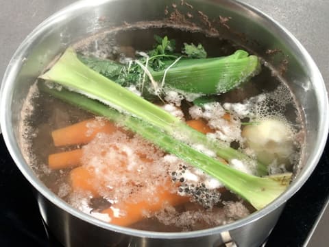 Ajout d'une branche de céleri dans l'eau frémissante dans le grand faitout qui contient les morceaux de viande, quatre carottes, un oignon, les grains de poivre et le bouquet garni