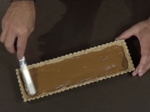 La ganache Dulcey est étalée à l'aide d'une petite spatule métallique coudée sur toute la surface du biscuit Muesli qui est dans le moule à tarte cannelé et rectangulaire