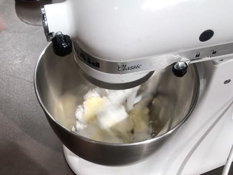 Ajout du beurre pommade sur le sucre glace dans la cuve du batteur électrique qui malaxe ces deux ingrédients avec l'accessoire feuille