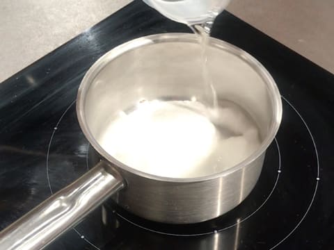 Un peu d'eau est versée sur le sucre en poudre dans la casserole qui est placée sur la plaque de cuisson