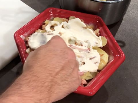 De la crème fraîche épaisse est étalée en une couche régulière sur les lardons et les oignons qui recouvrent les rondelles de pommes de terre dans le plat à gratin